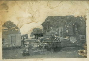 Weathered black and white 1950s photo of Chavis' house and store (left) and Jimmy's place (right). Three people stand in front of a vehicle between the two structures