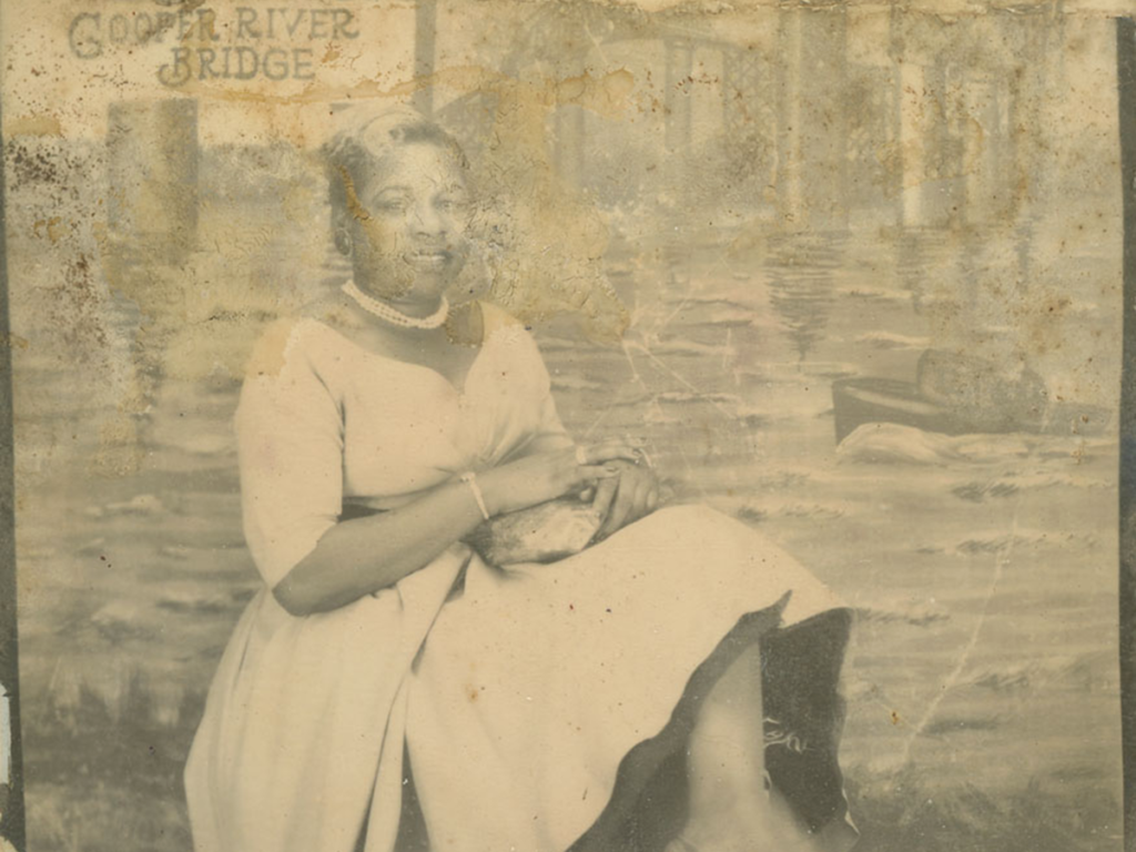 Sepia-toned photo fo a nicely dressed woman sitting in front of a mural that reads 